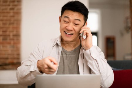 Foto de Retrato de un hombre asiático feliz trabajando desde casa, hablando por teléfono celular y usando un portátil, sentado en la sala de estar. Varón de mediana edad haciendo una llamada corporativa, teniendo conversación telefónica - Imagen libre de derechos