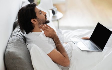 Téléchargez les photos : Jeune arabe malheureux en t-shirt blanc assis sur le lit, vérifie les amygdales, a appel vidéo sur ordinateur portable avec écran blanc, souffre de grippe et de froid dans l'intérieur de la chambre. Consultation en ligne, traitée à domicile - en image libre de droit