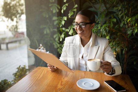 Téléchargez les photos : Heureuse femme d'affaires afro-américaine millénaire en costume blanc et verres à boire du café et des documents de lecture, menu sur tablette dans le bureau du café avec des plantes vertes à l'intérieur. Mode de vie, pause, entreprise - en image libre de droit