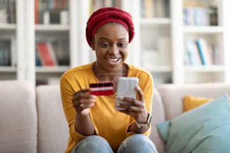 Photo for Excited happy pretty young african american woman wearing casual outfit and traditional turban sitting on couch, using cell phone and bank card at home, shopping while black friday, copy space - Royalty Free Image