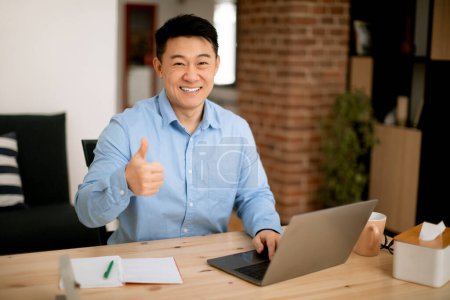 Foto de Me gusta. Feliz asiático de mediana edad hombre sentado en el lugar de trabajo en la oficina en casa, utilizando el ordenador portátil y mostrando el pulgar hacia arriba, disfrutando del trabajo remoto, mirando y sonriendo a la cámara - Imagen libre de derechos