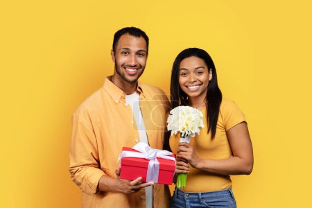 Photo for Cheerful millennial black guy gives bouquet and gift box to woman, congratulating with birthday, anniversary, Valentine day, isolated on yellow studio background. Love, romance, relationship - Royalty Free Image