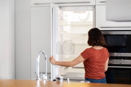 Photo for Housewife Looking Inside Of Empty Fridge While Standing In Kitchen, Hungry Young Woman Opening Refrigerator And Searching For Food, Having No Money To Buy Groceries, Rear View With Copy Space - Royalty Free Image