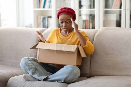 Téléchargez les photos : Déçu jolie jeune femme noire malheureuse portant turban africain traditionnel assis sur le canapé, tenant la boîte à papier, regardant à l'intérieur et gesticulant, client insatisfait déballer colis à la maison - en image libre de droit