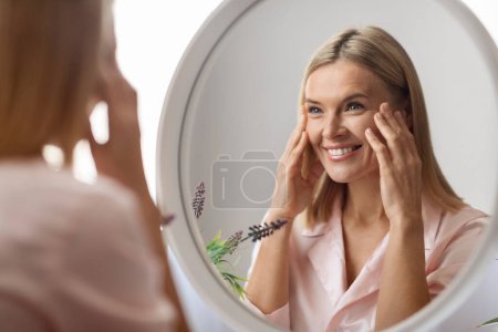 Foto de Concepto de belleza. Retrato de mujer madura atractiva mirando el espejo en el baño, hermosa señora de mediana edad tocando su piel de la cara impecable y sonriendo a la reflexión, enfoque selectivo - Imagen libre de derechos