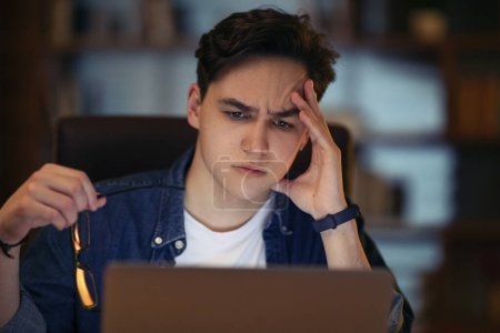 Photo for Closeup of concerned young guy freelancer sitting at workdesk at dark office late at night, looking at pc laptop screen, holding eyeglasses, touching his head, reading bad news, copy space - Royalty Free Image