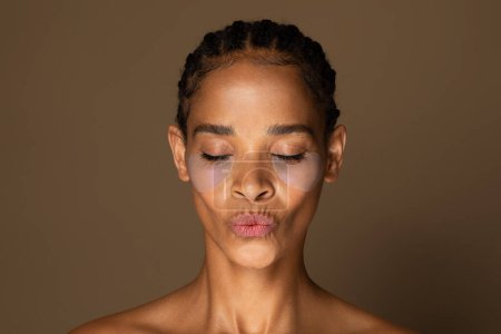 Téléchargez les photos : Femme noire d'âge moyen avec des taches d'hydrogel sous les yeux soufflant baiser, posant isolé sur fond brun. Publicité de produits cosmétiques, concept de beauté naturelle - en image libre de droit