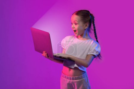 Téléchargez les photos : Incroyable excitée jolie fille d'âge scolaire élégant regardant l'écran brillant de l'ordinateur portable et criant, vérifiant le jeu vidéo le plus récent à couper le souffle, fond coloré avec effet de lumière néon - en image libre de droit
