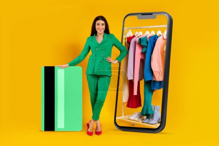 Photo for Happy lady standing near huge smartphone with clothing rail on display and giant credit card, using app or website, paying online, standing on yellow background - Royalty Free Image