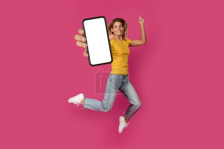 Photo for Cheerful middle aged european blonde lady jumping, freezes in air with smartphone with blank screen, has fun isolated on pink studio background. Recommendation app and device for work, great news - Royalty Free Image