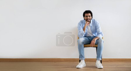 Foto de Sonriendo atractivo joven islámico masculino en casual y gafas se sienta en la silla, descansando, pensando, disfrutar del tiempo libre en fondo de pared gris en la habitación. Confort, alquilar o comprar casa, creando idea - Imagen libre de derechos