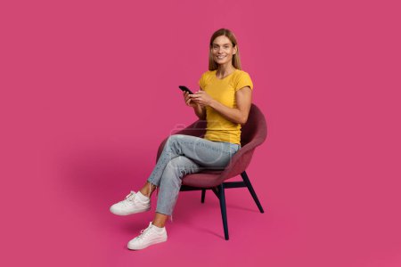 Photo for Glad cheerful middle aged european blonde lady sitting in chair, typing on smartphone isolated on pink studio background. App for work, business remotely with device, chat and communication - Royalty Free Image