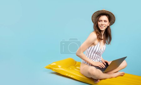 Téléchargez les photos : Femme heureuse en maillot de bain et chapeau de paille assis sur matelas gonflable de natation et en utilisant ordinateur portable PC isolé sur fond bleu, panorama avec espace de copie - en image libre de droit