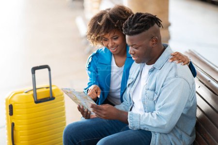 Téléchargez les photos : Joyeux jeunes conjoints noirs vérifiant la route de voyage avec la carte de la ville tout en étant assis sur le banc à la gare, heureux couple afro-américain Choisir une destination de voyage ensemble en attendant le train - en image libre de droit