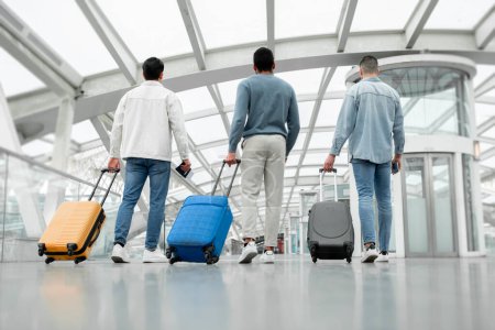 Téléchargez les photos : Billets de voyage pas chers. Vue arrière de trois hommes amis groupe marchant avec des valises posant de retour à la caméra dans le terminal de l'aéroport à l'intérieur. Prise de vue pleine longueur de passagers méconnaissables - en image libre de droit