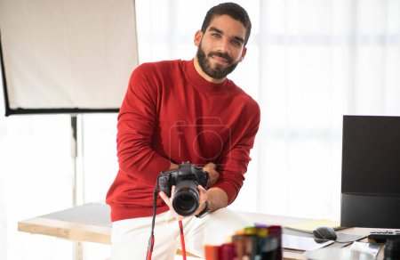 Téléchargez les photos : Positif souriant beau jeune homme hispanique en tenue décontractée élégant photographe professionnel posant à son studio photo de travail, assis sur le bureau avec PC, tenant un appareil photo numérique, espace de copie - en image libre de droit