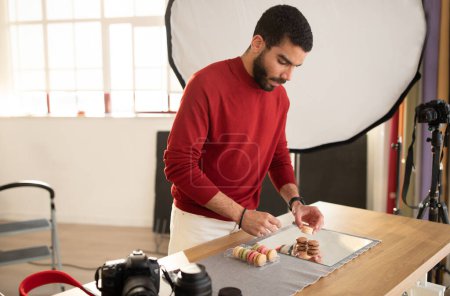 Photo for Photographer take photo of sweets in photo studio, setting macaroons on mirror board. Concentrated bearded handsome young man photographing colorful macaroons dessert, copy space - Royalty Free Image