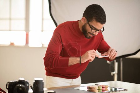 Photo for Handsome young hispanic bearded man wearing casual outfit and eyewear photographing delicious tasty sweet dessert colorful macaroons on mirror board, using smartphone, photo studio interior - Royalty Free Image