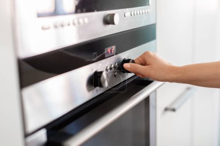 Unrecognizable Woman Using Electic Oven In Kitchen, Adjusting Temperature With Hand, Young Housewife Turning Knob, Selecting Burner Cooking Mode While Preparing Food At Home, Closeup Shot