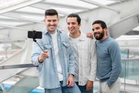 Téléchargez les photos : Groupe de trois heureux amis masculins voyageant et faisant Selfie sur téléphone portable s'amuser en attendant le vol dans l'aéroport moderne à l'intérieur. Blog Voyage vacances et Gadgets Concept - en image libre de droit