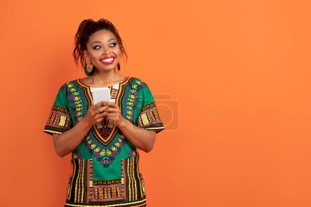 Photo for Dreamy curious happy pretty young black woman wearing african clothing and bright makeup with phone in her hands looking at copy space and smiling, isolated on orange background. Online shopping - Royalty Free Image