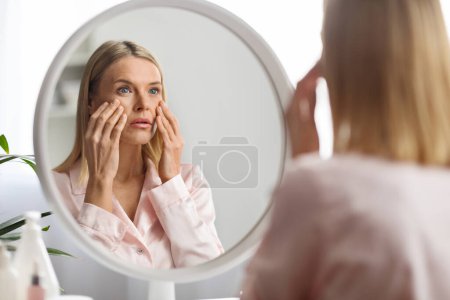 Hautprobleme Konzept. Verstörte Frau mittleren Alters beim Anblick von Spiegel und anrührendem Gesicht, verwirrte ältere Dame bemerkte Falten und Augenringe, unzufrieden mit der Hautpflege-Routine, selektiver Fokus