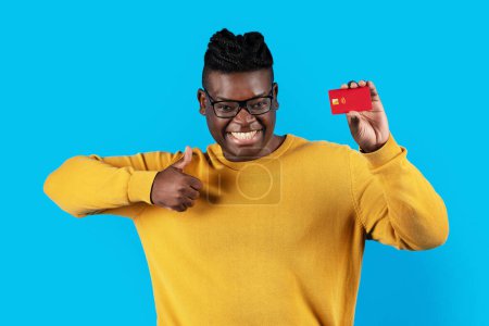 Photo for Handsome Young Black Man Pointing At Credit Card In His Hand And Looking At Camera, Happy Millennial African American Male Recommending Bank Services While Standing On Blue Studio Background - Royalty Free Image