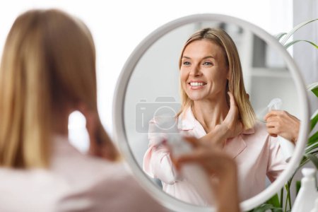 Photo for Beautiful Middle Aged Woman Applying Moisturising Hair Spray On Ends While Sitting Near Mirror At Home, Attractive Mature Lady Enjoying New Hairstyle Cosmetics And Smiling To Her Reflection - Royalty Free Image