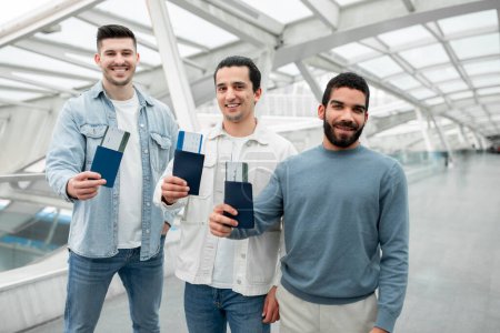 Foto de Grupo de tres hombres sonrientes mostrando tarjetas de embarque y pasaportes mirando la cámara de pie en el aeropuerto moderno en interiores. Amigos masculinos alegres que viajan juntos Publicidad Oferta de viaje - Imagen libre de derechos
