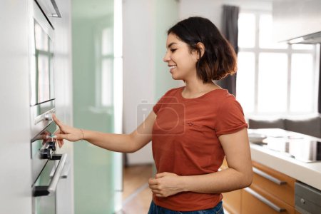 Téléchargez les photos : Heureuse jeune femme au foyer arabe utilisant une cuisinière moderne tout en préparant la nourriture à la maison, Souriante femme du Moyen-Orient allumant le four électrique à l'intérieur de la cuisine, Appuyant sur le bouton avec le doigt, Espace de copie - en image libre de droit