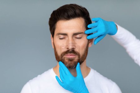 Téléchargez les photos : Portrait d'un homme d'âge moyen barbu se préparant pour les procédures de chirurgie plastique, médecin touchant la peau du visage masculin, isolé sur fond gris, espace de copie - en image libre de droit