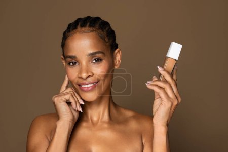 Téléchargez les photos : Portrait de belle femme noire d'âge moyen avec une peau propre et fraîche, tenant la bouteille de maquillage de fond de teint et souriant à la caméra, posant sur fond brun. Recommandation de produit de maquillage - en image libre de droit