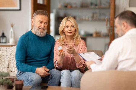 Photo for Mature Couple Consulting With Health Advisor Holding Vitamins Pills Sitting Indoor. Spouses Talking With Doctor Or Nutrition Specialist About Medical Supplements And Treatment. Selective Focus - Royalty Free Image