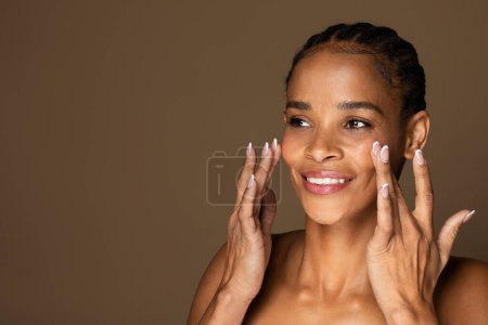 Photo for Skincare concept. Portrait of beautiful black woman with smooth skin touching face, applying cream and looking aside at free space, standing isolated over brown background - Royalty Free Image