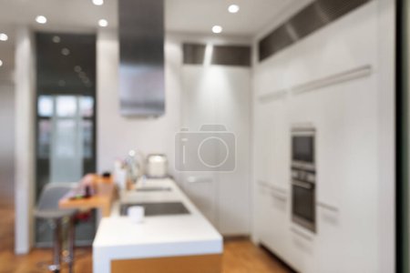Téléchargez les photos : Vue déconcentrée de l'intérieur de la cuisine élégante dans un appartement moderne, design minimaliste contemporain avec armoires blanches, appareils de cuisine, chaises et comptoir spacieux, image floue - en image libre de droit