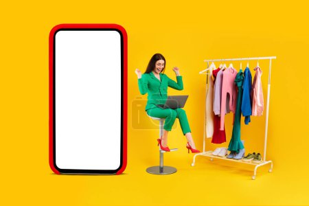 Photo for Happy lady shopaholic sitting near huge phone and clothing rail, using laptop and gesturing yes, approving online store, yellow background. Shopping concept. Mockup - Royalty Free Image