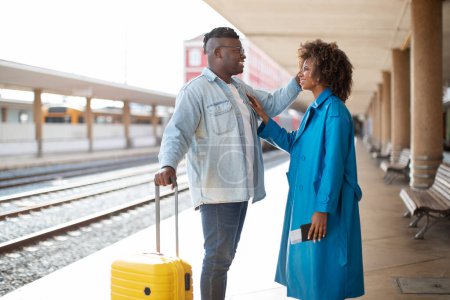 Foto de Viaje de luna de miel. Cónyuges negros románticos felices que esperan el tren en la estación de tren, pareja afroamericana encantadora que está parada en la plataforma con la maleta y abrazando, disfrutando de viajar juntos - Imagen libre de derechos
