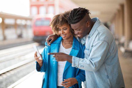Foto de Turistas negros se unen usando Smartphone mientras esperan tren en la estación de tren, cónyuges afroamericanos felices de pie en la plataforma y reservar boletos de viaje en aplicación móvil, espacio libre - Imagen libre de derechos