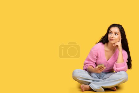 Téléchargez les photos : Réfléchie millénariste arabe pieds nus dame avec smartphone se trouve sur le sol et regarde l'espace de copie isolé sur fond jaune studio. Vente, maquette, annonce de vacances et offre, créer une idée - en image libre de droit