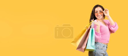 Photo for Cheerful millennial middle eastern lady in sunglasses with many bags of purchases enjoy shopping and looks at empty space isolated on yellow studio background. Relax, lifestyle, fashion, sale - Royalty Free Image