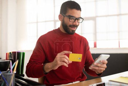 Foto de Sonriente joven apuesto hombre barbudo hispano en gafas de uso casual sentado en el escritorio en la oficina, amarillo usando tarjeta bancaria y teléfono inteligente, banca de hombre de negocios, compras en línea, espacio de copia - Imagen libre de derechos