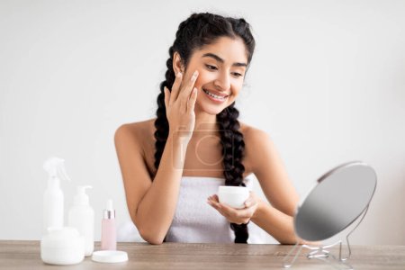 Photo for Skin Care Concept. Smiling Beautiful Indian Woman Applying Moisturising Face Cream While Sitting In Front Of Mirror At Home, Happy Eastern Female Making Beauty Skincare Routine, Copy Space - Royalty Free Image