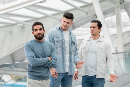 Photo for Three Male Friends Having Quarrel Traveling Together Standing In Modern Airport Terminal Indoor. Man Sulking Crossing Hands Having Conflict With Travel Companions. Communication Problems - Royalty Free Image