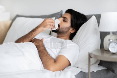 Photo for Sad young middle eastern guy in white t-shirt lies on bed, blows nose in napkin, sick and suffers from flu and cold in bedroom interior. Treated at home, health care, allergy - Royalty Free Image