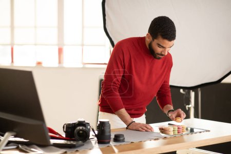 Photo for Graphic designer handsome young hispanic man in stylish casual outfit working on food advertisement at modern spacious office, taking photo of dessert colorful macaroons, copy space - Royalty Free Image