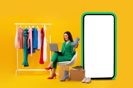 Photo for Stylist woman using laptop and holding elegant shoe, sitting near clothing rail and big phone with blank screen, selling outfits over internet on yellow studio background, mockup - Royalty Free Image