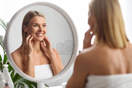 Téléchargez les photos : Concept de beauté. Souriant attrayant moyen âge femme regardant son reflet dans le miroir à la maison, belle femme mûre enveloppée dans une serviette de bain bénéficiant de son apparence, foyer sélectif - en image libre de droit