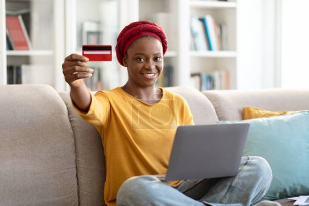 Foto de Feliz sonrisa hermosa mujer negra joven en turbante rojo usando el ordenador portátil, mostrando la tarjeta bancaria en la cámara, sentado en el sofá en casa, pedir comida en línea, pagar por los bienes y servicios en Internet, espacio para copiar - Imagen libre de derechos