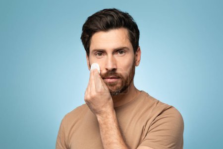 Photo for Handsome bearded man applying cleansing lotion on his face, using cotton pad and looking at camera, isolated over blue studio background. Beauty and skin care concept - Royalty Free Image