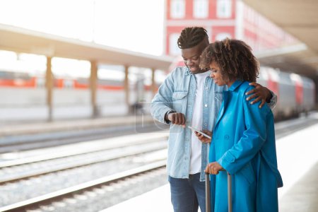 Foto de Cónyuges negros felices esperando el tren en la estación de tren, sonriendo hombre y mujer afroamericanos viajando juntos, parados en la plataforma y comprobando la hora de salida en el reloj de pulsera, espacio de copia - Imagen libre de derechos
